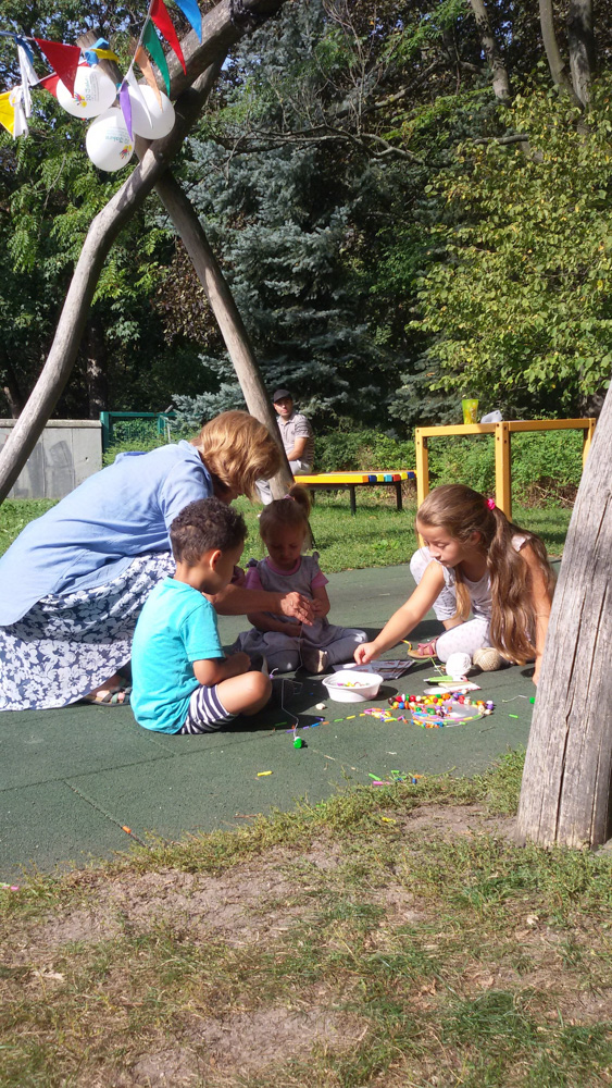 Foto: Kinder und Erzieherin sitzen auf der Erde und fertigen Perlenketten