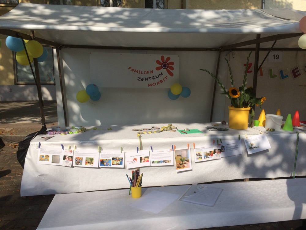Foto: der Stand vom Familienzentrum mit aufgehängten Fotos an Forderseite und mit Logo-Banner im Hintergrund. Davor ein Tisch mit Buntstiften im Becher und Papier zum bemalen.