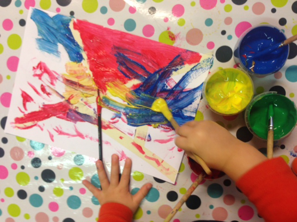 Foto: Tuschezeichnung, Tuschefarbtöpfchen blau, gelb und grün und Kinderhände bzw. -hand mit Pinsel beim zeichnen. Draufsicht von oben.