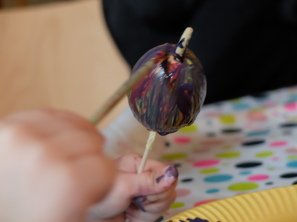 Foto: ein ausgeblasenes Ei auf Holzstil von linker Kinderhand gehalten und mit rechter Kinderhand mit Pinsel bemalt.