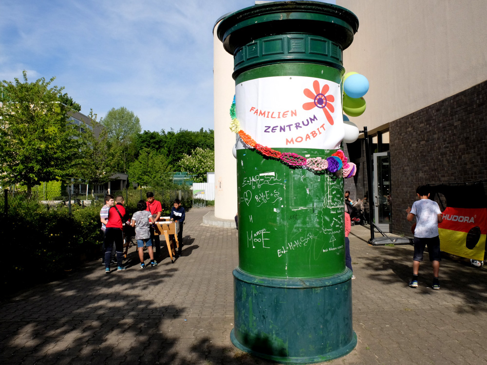 Foto: grüne Litfaßsäule mit Luftballons und Girlanden sowie dem Schriftzug bzw. Logo Familien Zentrum Moabit. Kinder spielen im Hintergrund.