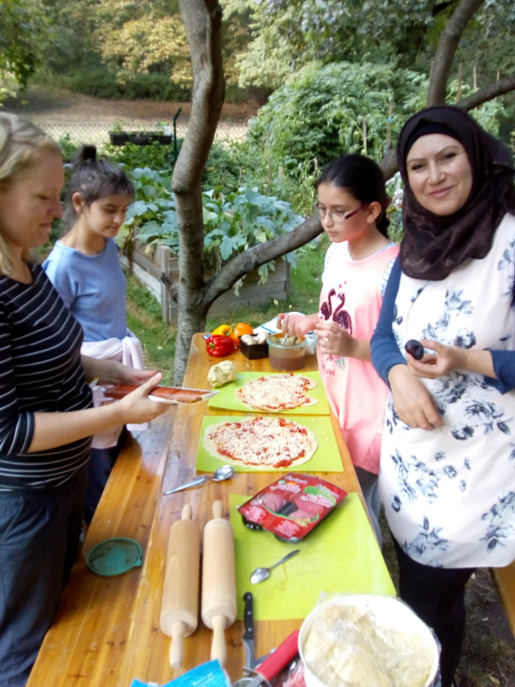 Foto: Frau mit Kopftuch und ohne und 2 Mädchen belegen Pizzateig na einem langen Tisch.