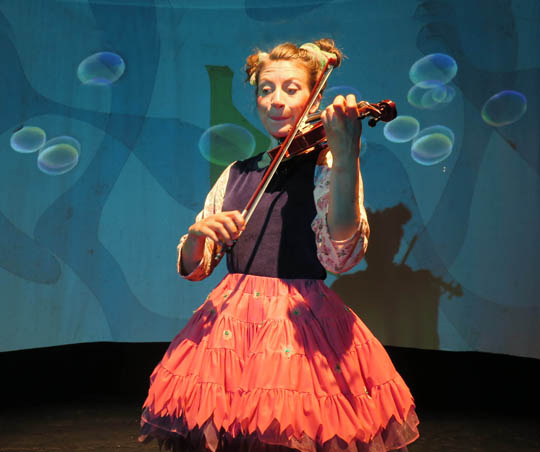 Foto: Frau mit rotem Rock spielt Violine auf einer Bühne mit blauem Hintergrund