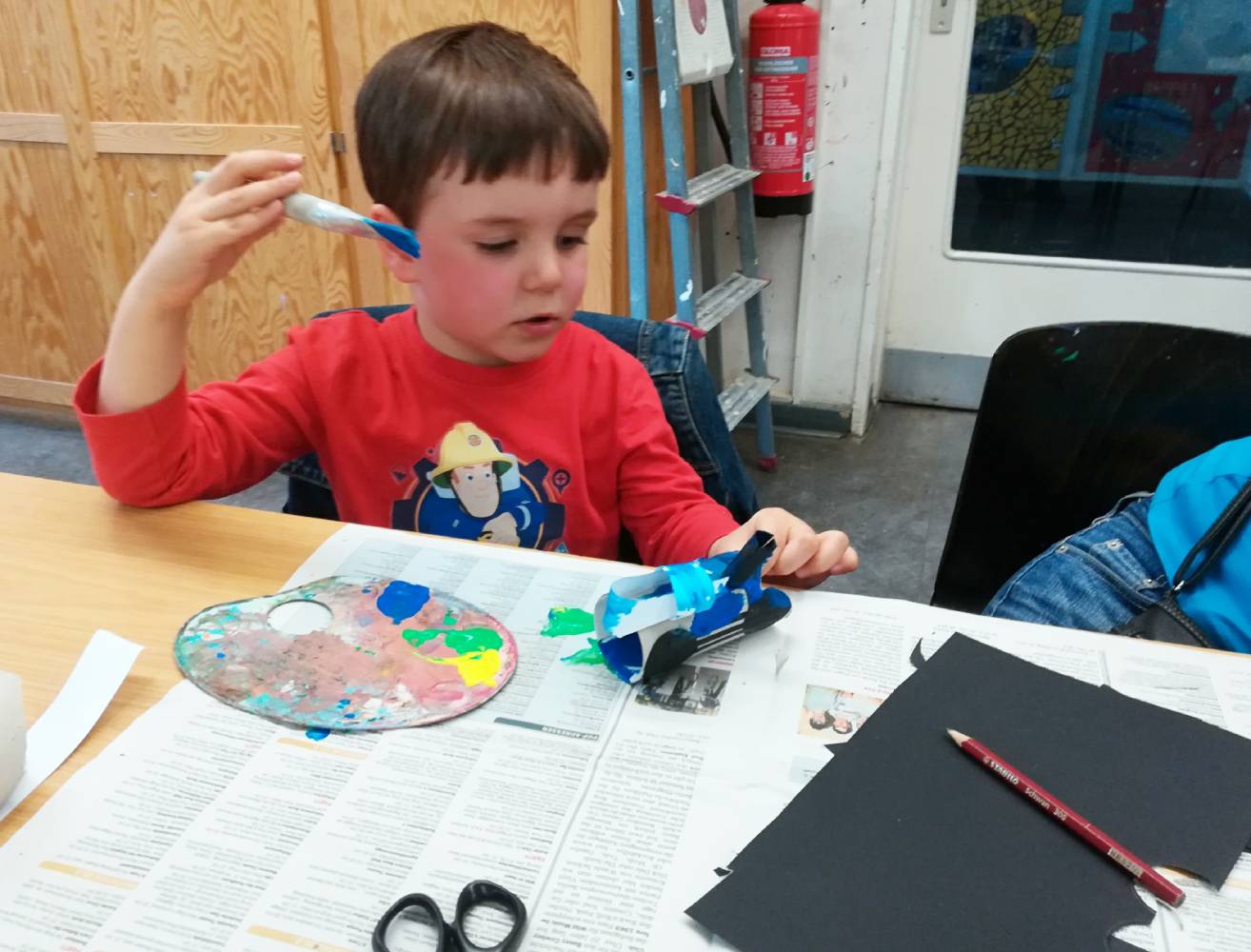 Foto: Kind an Tisch sitzend hält blauen Pinsel in der rechten Hand und malt Klopapierrolle an