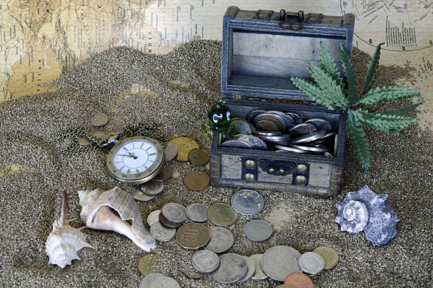 Foto: oben rechts eine kleine Schatztruhe auf Sand stehend, davor Münzen, Muscheln und eine Uhr im Hintergrund eine Schatzkarte