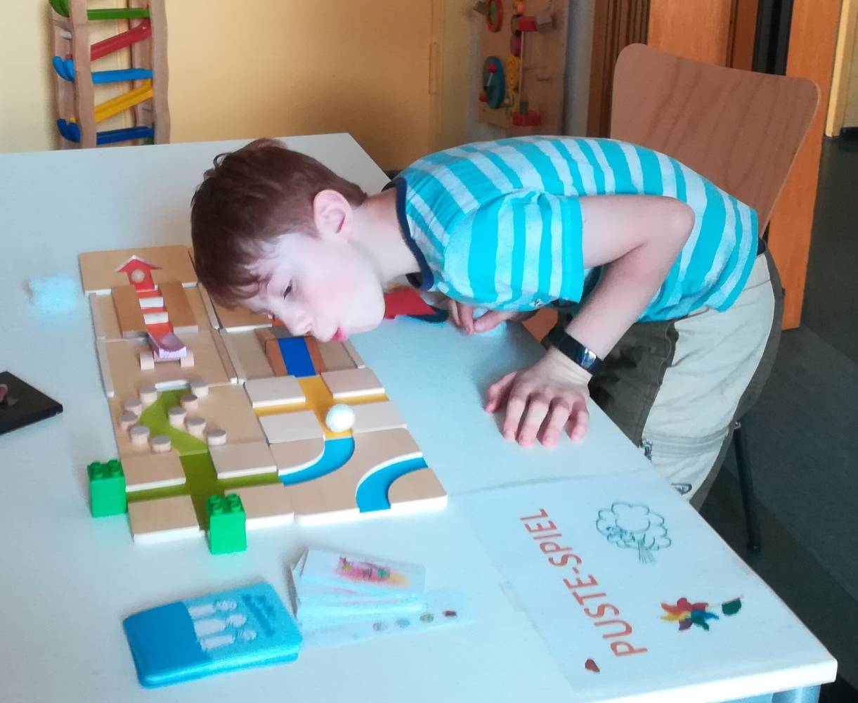 Foto: Ein Tisch mit einem Spiel drauf, rechts im Bild ein Junge, der auf das Spiel pustet