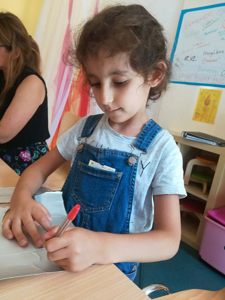 Foto: Mädchen sitzt am Tisch und malt mit einem rotem Stift auf silbernen Karton