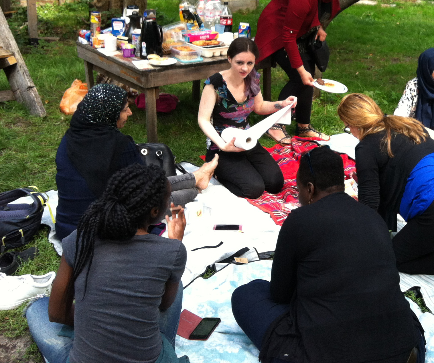 Foto: Decken auf Wiese mit fünf Frauen, die auf dieser sitzen. Im Hintergrund ein Holztisch mit Essen