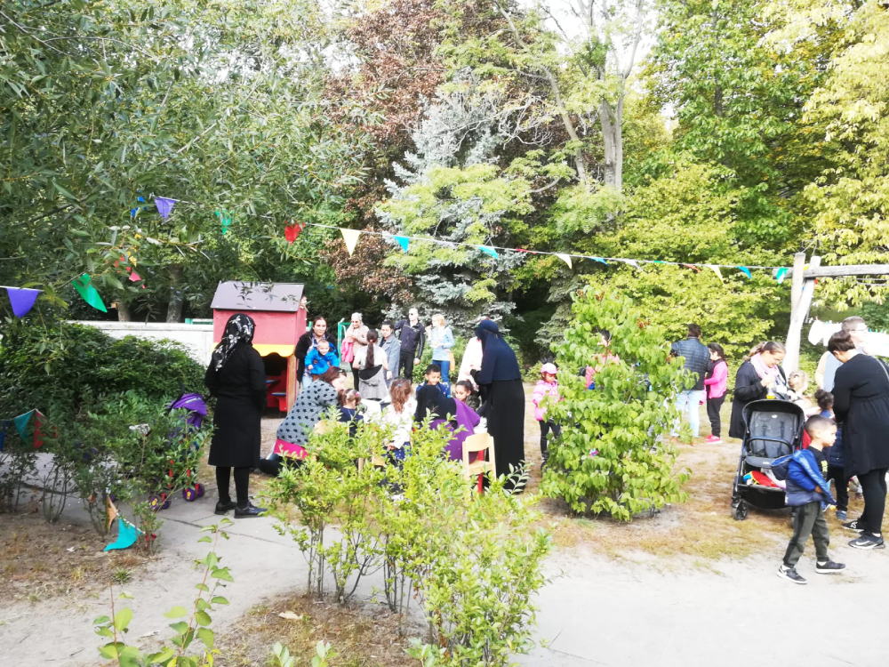 Foto: Draußen, Bäume und Sträucher, dazwischen Girladen. In der Mitte stehen und sitzen viele Familien mit kleinen Kindern