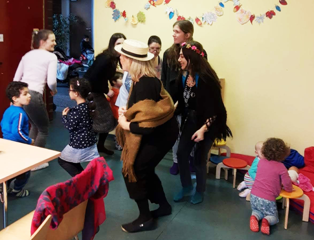 Foto: Erwachsene und Kinder Tanzen links im Bild, rechts spielen zwei Kleinkinder