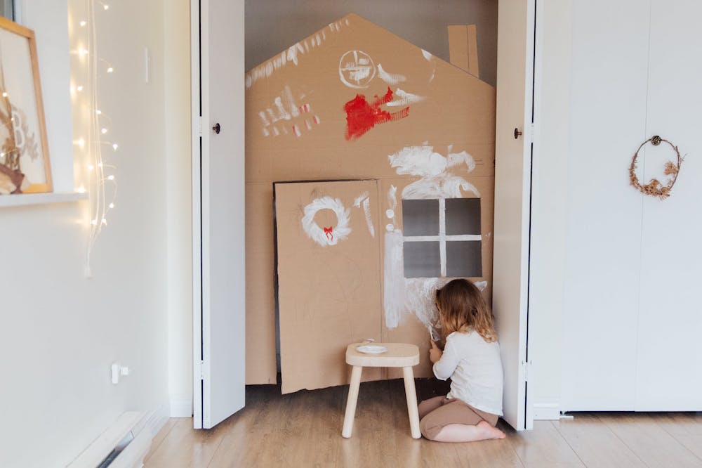 Foto: Kind vor Schrank, in den ein Papphaus steht. Kind malt Papphaus weiß an.