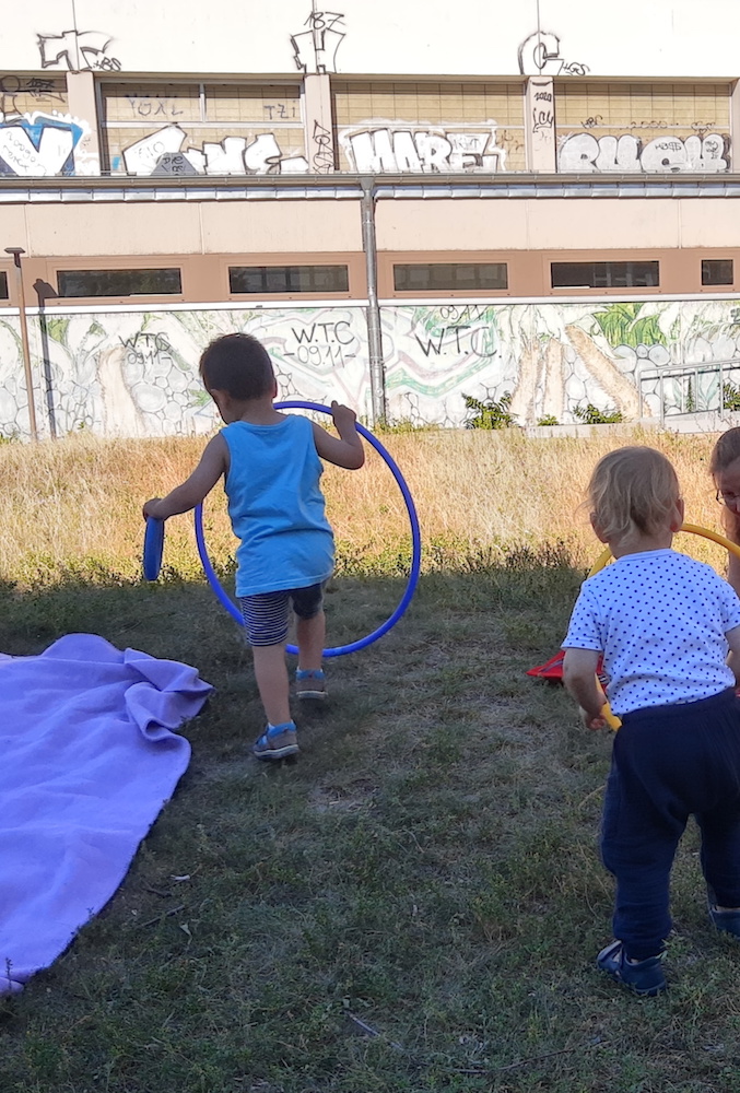 Foto: zwei Kinder von hinten, die eine Wiese entlanggehen. Beide halten große Hula-Hoop-Reifen.