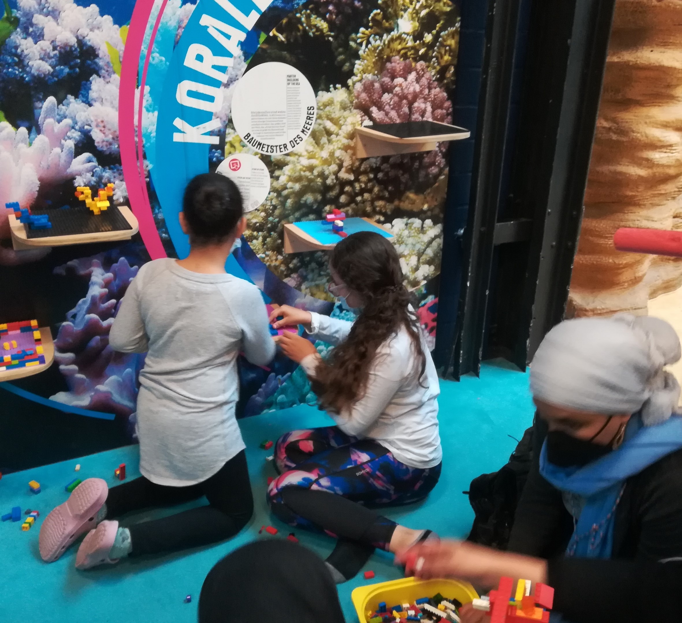 Foto: zwei Kinder vor dem Bild eines Korallenriffs die mit kleinen Legosteinen etwas bauen, rechts unten eine Frau mit einer Kiste Legosteinen
