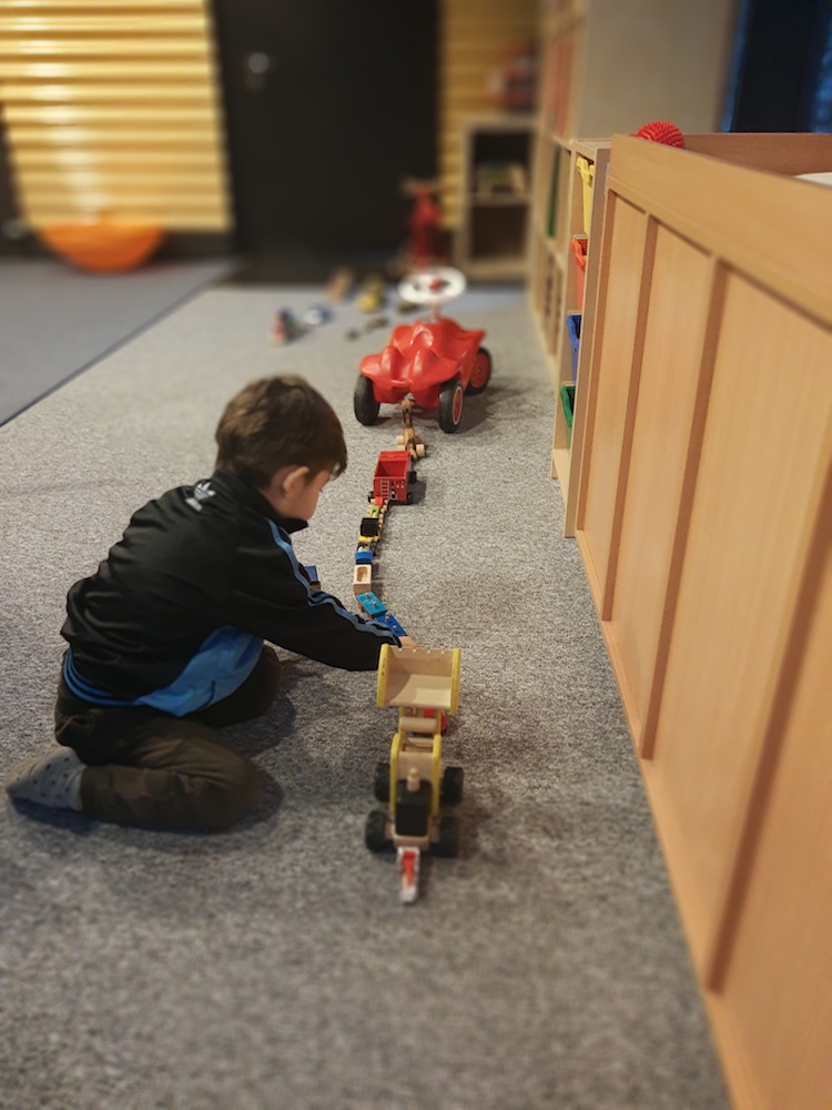 Foto: Links im Bild sitzt ein Kind im Bild, dass mit Holzspielzeugautos und -zügen, rechts im Bild, spielt.
