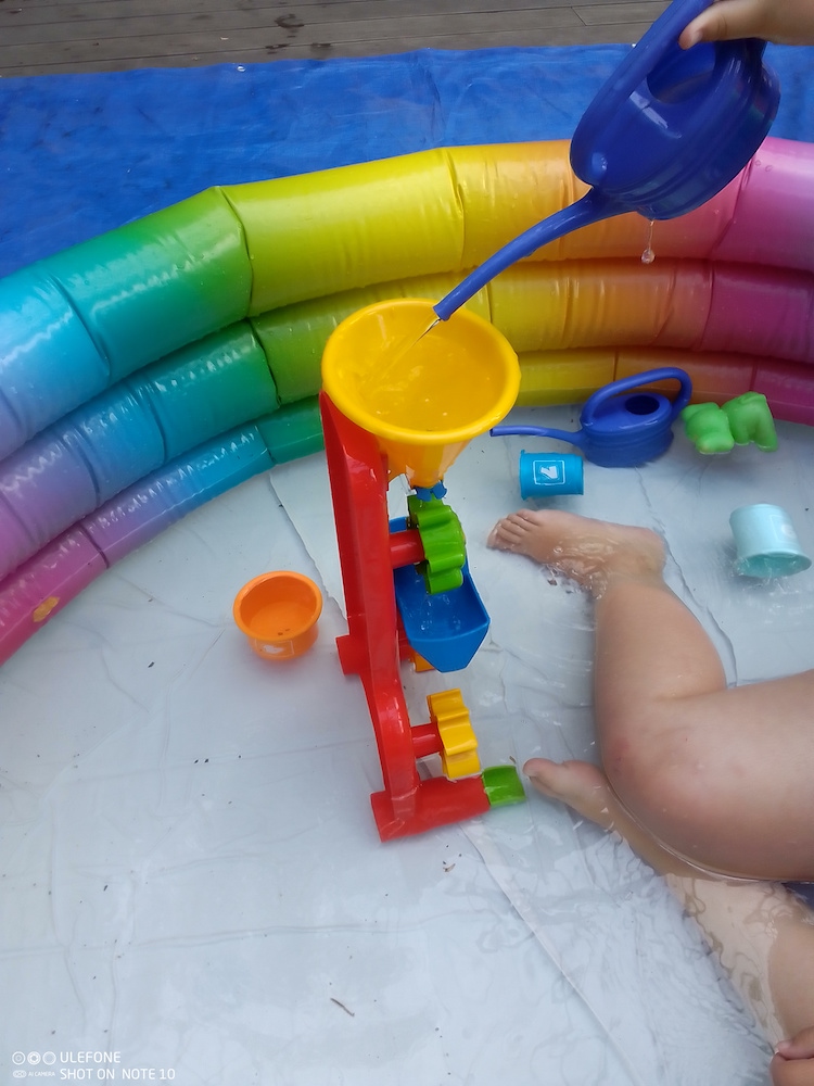 Foto: Rand von Plantschbecken, rechts Kinderbeine und Wasserspielzeug, eine blaube Gießkanne von oben